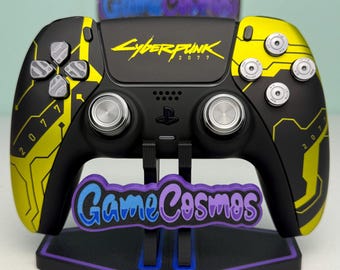 May include: A black and yellow wireless video game controller featuring Cyberpunk 2077 branding, metallic silver buttons, and analogue sticks. The controller is mounted on a black stand with a purple and blue GameCosmos logo attached to the front.