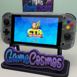 Könnte beinhalten: Eine transparente Handheld-Spielkonsole, die das Spiel "Crash Team Racing Nitro-Fueled" anzeigt. Die Konsole steht auf einem schwarzen Ständer mit einem lila-blauen "Game Cosmos"-Logo. Ein passendes Logo ist im Hintergrund sichtbar.