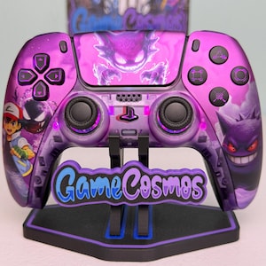 May include: A custom purple video game controller featuring a graphic of Gengar and Ash Ketchum. The controller has black analog sticks, buttons, and a central PlayStation logo. A black stand with the text GameCosmos is positioned underneath.