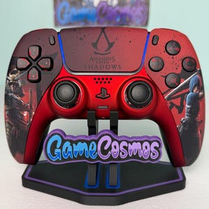 Op de afbeelding: Een rode en zwarte gamecontroller met de tekst "ASSASSINS CREED SHADOWS". De controller staat op een zwarte standaard met de tekst "Game Cosmos". De controller heeft zwarte knoppen en zwarte joysticks.