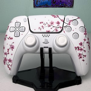 May include: A white wireless video game controller featuring a pink cherry blossom branch pattern across the front shell, including two white analog sticks, a central PlayStation logo, and a black plastic display stand underneath.