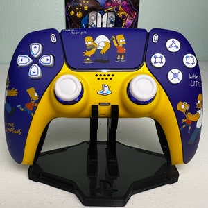 Könnte beinhalten: Ein gelber und blauer, individuell gestalteter Playstation 5-Controller mit Simpsons-Charakteren und dem Text "Mmm... floor pie" und "Why you little..." auf dem Controller. Der Controller steht auf einem schwarzen Ständer.