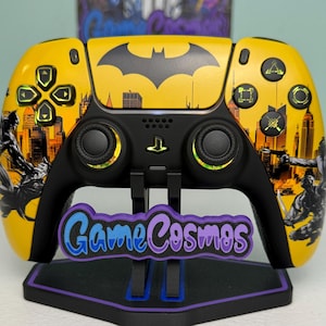 May include: A yellow and black wireless video game controller featuring a Batman logo and cityscape graphic. The controller has black analog sticks, a central PlayStation button, and action buttons with yellow accents. The text GameCosmos is visible.
