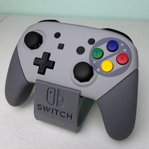 Puede incluir: Un mando gris de Nintendo Switch con un dise&ntilde;o personalizado de Super Nintendo. El mando tiene un esquema de color gris y negro con los botones cl&aacute;sicos de Super Nintendo: A, B, X, Y y Start. El mando est&aacute; sentado en un soporte gris con el logotipo de Nintendo Switch.