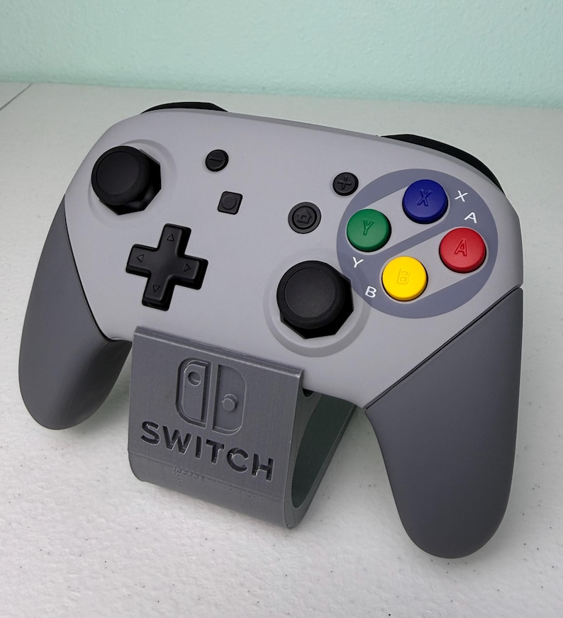 Puede incluir: Un mando gris de Nintendo Switch con un dise&ntilde;o personalizado de Super Nintendo. El mando tiene un esquema de color gris y negro con los botones cl&aacute;sicos de Super Nintendo: A, B, X, Y y Start. El mando est&aacute; sentado en un soporte gris con el logotipo de Nintendo Switch.