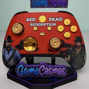 May include: A red video game controller featuring the text Red Dead Redemption, gold bullet-shaped buttons, and gold joysticks. The controller displays illustrations of two figures in cowboy attire and is mounted on a black stand with the text GameCosmos.