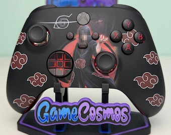 May include: A black wireless video game controller featuring a graphic of a character in a dark cloak with red clouds. The controller has red backlit buttons and a D-pad. It rests on a black stand with the purple and blue text GameCosmos.