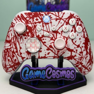 May include: White Xbox controller with a red splatter design, resting on a black stand with the text "GameCosmos" in blue and purple. The controller has white buttons and a glowing blue Xbox logo.
