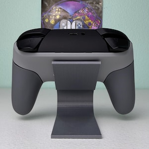 Puede incluir: Un soporte gris para mando de videojuegos con un mando gris en la parte superior. El mando tiene botones negros y una parte superior negra.