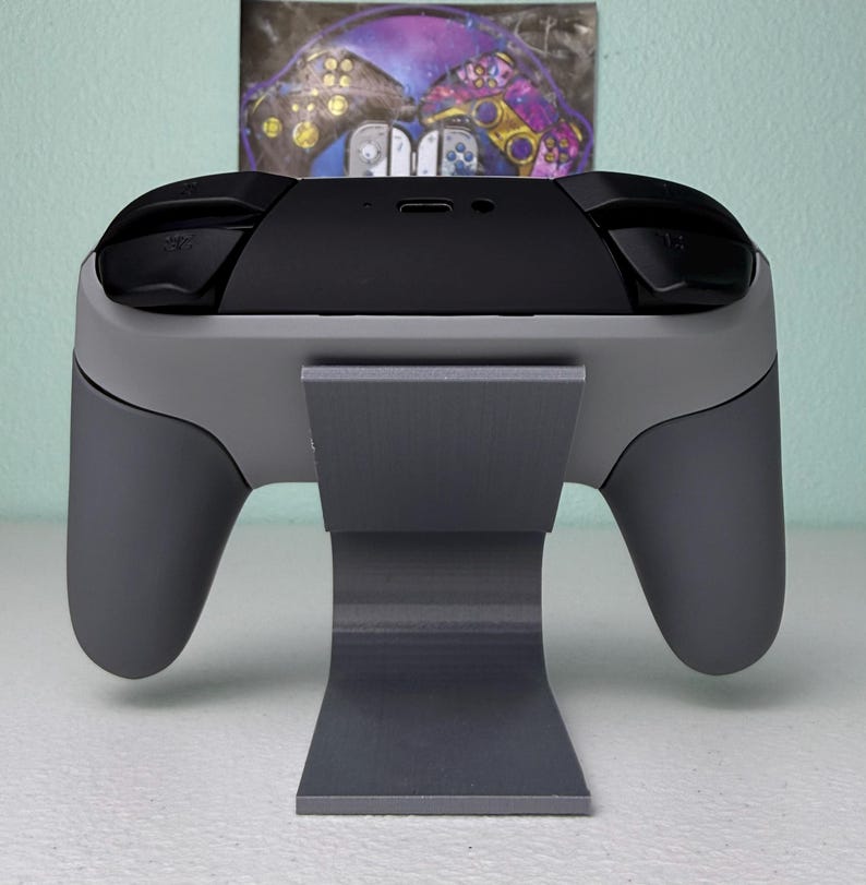 Puede incluir: Un soporte gris para mando de videojuegos con un mando gris en la parte superior. El mando tiene botones negros y una parte superior negra.