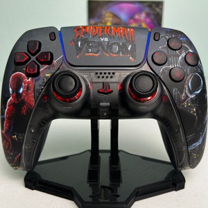 May include: Black and red Spider-Man vs. Venom PlayStation 5 controller. The controller features Spider-Man and Venom graphics, with the text "Spider-Man vs. Venom" across the top. The controller is on a black stand.