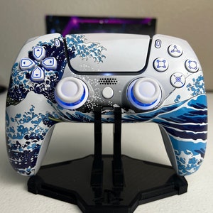 May include: A white PlayStation 5 DualSense controller with a blue and white wave design. The controller is on a black stand.