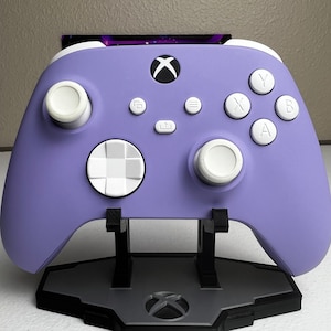 Puede incluir: Un mando Xbox morado con botones blancos y un pad direccional blanco. El mando está en un soporte negro con un logotipo de Xbox.