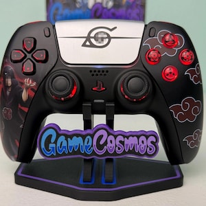May include: A black wireless video game controller featuring red accents, a silver center panel with a leaf symbol, and red cloud graphics. The controller includes red button icons and a character illustration on the left side, displayed on a black stand.