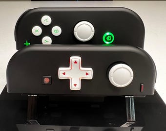 Może przedstawiać: Dwa czarne kontrolery Nintendo Switch Joy-Con zamontowane na podstawce. Górny kontroler posiada białe przyciski A, B, X, Y, biały joystick oraz świecący na zielono przycisk home. Dolny kontroler ma biały krzyżak kierunkowy z czerwonymi strzałkami.