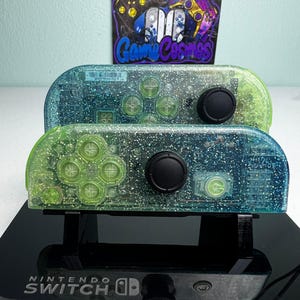 May include: Two Nintendo Switch Joy-Con controllers featuring a clear, glitter-infused shell with a blue-to-green gradient. The controllers have black analog sticks and light green buttons, displayed on a black stand with the Nintendo Switch logo.