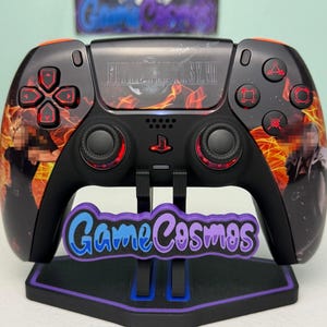 May include: A black wireless video game controller featuring Final Fantasy VII graphics with orange flames, red button accents, and the text GameCosmos on a purple and blue stand. The controller has two analog sticks and a central PlayStation logo.