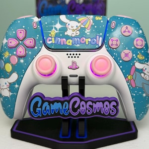 May include: A custom video game controller featuring a teal blue shell with a Cinnamoroll character print, colorful balloons, and stars. The controller has pink thumbsticks, a white center panel, and a black stand with the text GameCosmos.