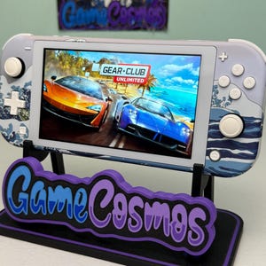 May include: A handheld gaming console with a screen displaying a racing game. The console is white and gray with a wave pattern design. It rests on a black and purple stand with the text "Game Cosmos".