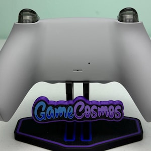 Puede incluir: Un mando de juego gris claro sobre un soporte negro y morado. El soporte presenta el texto "GameCosmos" en azul y morado. El mando tiene dos botones transparentes y redondeados en la parte superior.