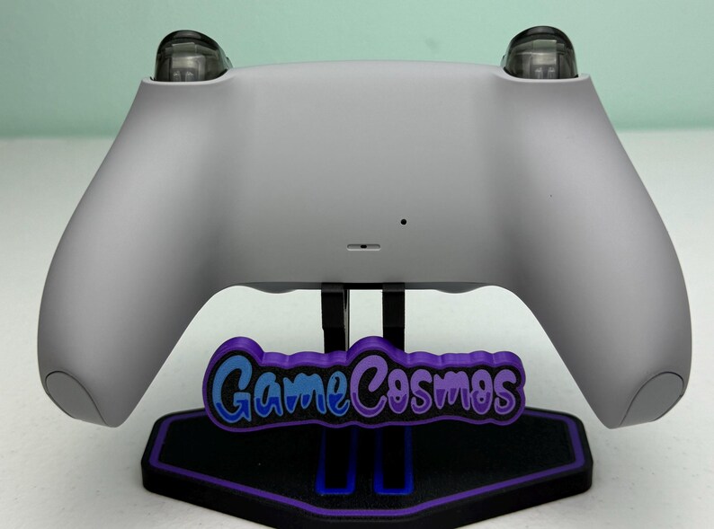 Puede incluir: Un mando de juego gris claro sobre un soporte negro y morado. El soporte presenta el texto "GameCosmos" en azul y morado. El mando tiene dos botones transparentes y redondeados en la parte superior.