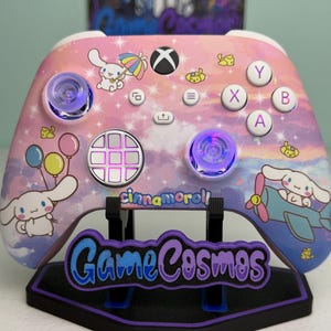 May include: A wireless video game controller featuring a pastel pink, purple, and blue sky-themed shell with Cinnamoroll character graphics, including a character with an umbrella and a plane. The controller has white buttons, glowing purple thumbsticks, and the text Cinnamoroll printed on the front surface.