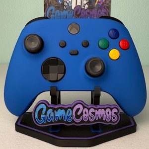 May include: A blue Xbox controller with black detailing, displayed on a stand. The stand is black with a purple and blue glitter design that reads "Game Cosmos". The controller has coloured buttons.