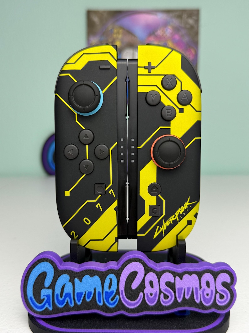 Puede incluir: Mandos Joy-Con de Nintendo Switch con tem&aacute;tica Cyberpunk 2077 en negro y amarillo. Los mandos tienen un dise&ntilde;o de placa de circuito con el texto "2077" y "Cyberpunk". Los mandos est&aacute;n sobre un soporte con el texto "Game Cosmos".