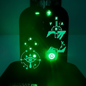 Può includere: Due controller di gioco neri su una base Nintendo Switch con design luminosi di colore verde. Il controller sinistro mostra il simbolo dell'occhio Sheikah, mentre quello destro presenta una grafica a spada con i pulsanti X, Y, A e B.