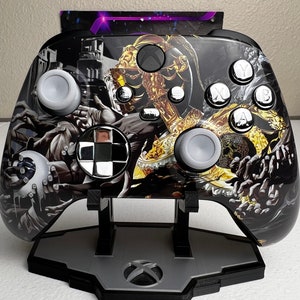 May include: A black and gold Xbox controller with a custom design featuring a skull and other imagery. The controller is on a black stand.