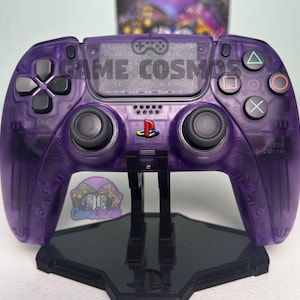 May include: A purple translucent PlayStation 5 DualSense controller with a black stand. The controller has a textured surface and the text "GAME COSMOS" is printed on the front.
