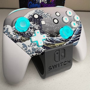 Puede incluir: Un mando blanco de Nintendo Switch con un dise&ntilde;o azul y blanco de la Gran Ola de Kanagawa. El mando est&aacute; en un soporte negro con la palabra "SWITCH" impresa en &eacute;l.
