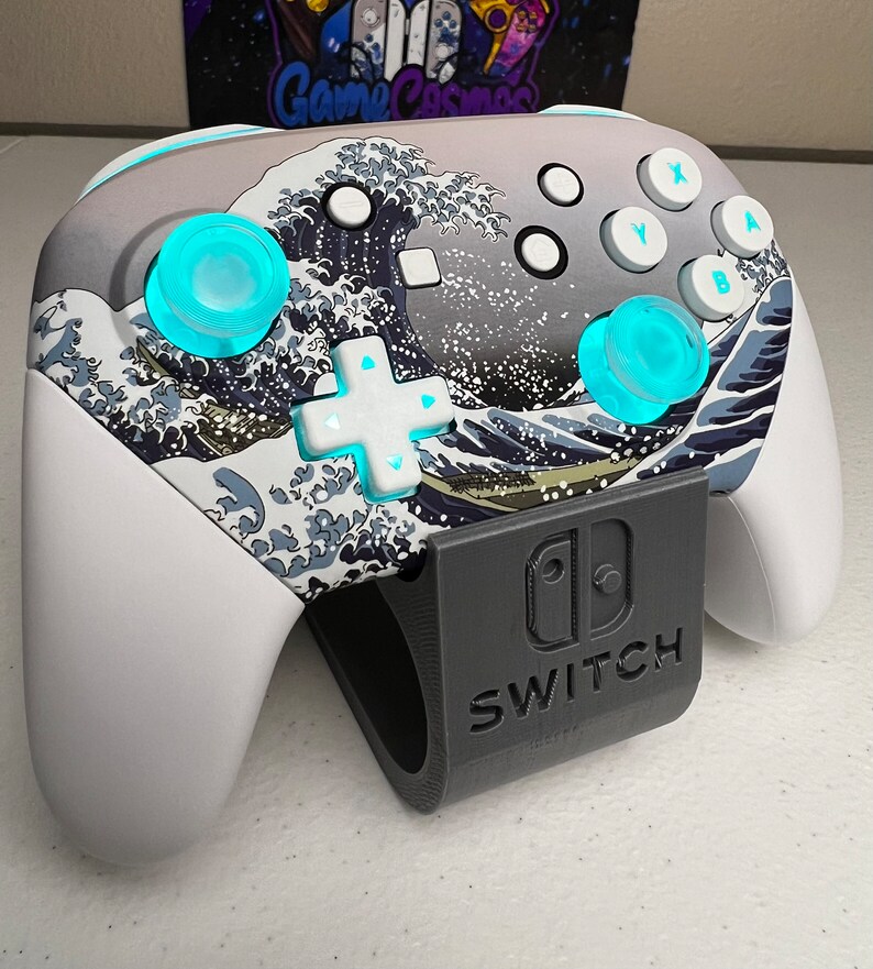 Puede incluir: Un mando blanco de Nintendo Switch con un dise&ntilde;o azul y blanco de la Gran Ola de Kanagawa. El mando est&aacute; en un soporte negro con la palabra "SWITCH" impresa en &eacute;l.