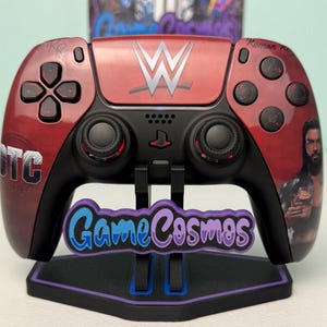 Può includere: Un controller PlayStation 5 rosso e nero personalizzato con il logo WWE e un ritratto di Roman Reigns. Il controller è su un supporto con il testo "Game Cosmos" in blu e viola.