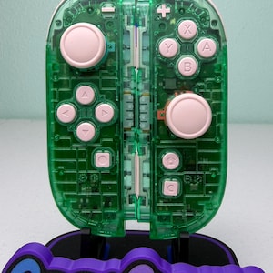 May include: A transparent green video game controller with pink buttons and a black and purple base that reads "Game Cosmos". The controller has a clear design, revealing internal components.