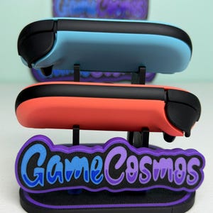 Pu&ograve; includere: Un espositore che contiene due controller di gioco, uno azzurro e uno corallo, con dettagli neri. L'espositore presenta il testo "Game Cosmos" in blu e viola, su una base nera e viola.