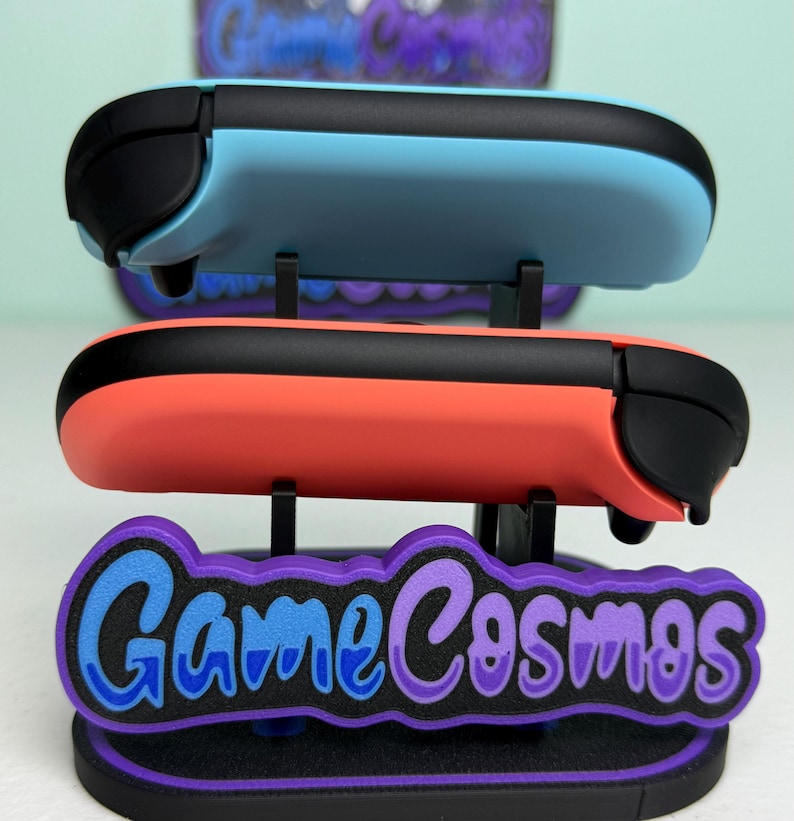 Pu&ograve; includere: Un espositore che contiene due controller di gioco, uno azzurro e uno corallo, con dettagli neri. L'espositore presenta il testo "Game Cosmos" in blu e viola, su una base nera e viola.