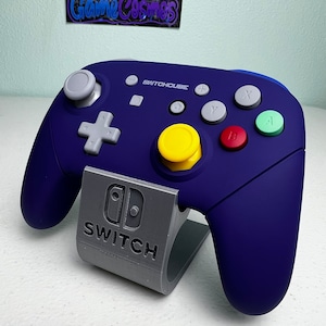 May include: A blue and gray Nintendo Switch controller with the text "SWITCHCUBE" on the front. The controller is sitting on a gray stand with the Nintendo Switch logo and the word "SWITCH" printed on it.