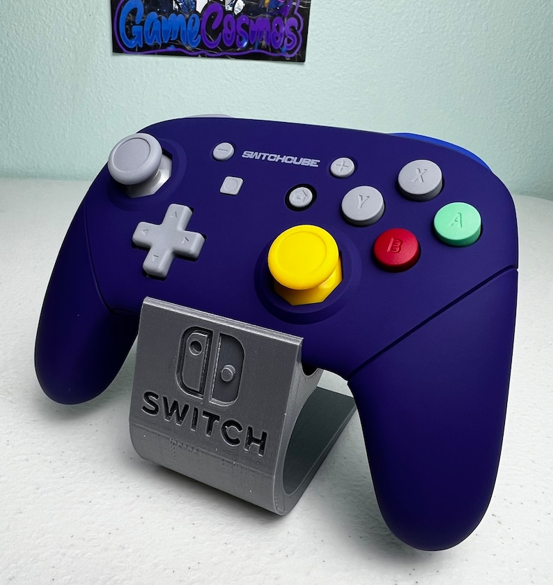 May include: A blue and gray Nintendo Switch controller with the text "SWITCHCUBE" on the front. The controller is sitting on a gray stand with the Nintendo Switch logo and the word "SWITCH" printed on it.