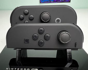 Może przedstawiać: Dwa matowoczarne kontrolery Joy-Con do konsoli Nintendo Switch umieszczone na czarnym stojaku ekspozycyjnym. Górny kontroler posiada przyciski A, B, X, Y oraz joystick. Dolny kontroler ma krzyżak kierunkowy i joystick. Podstawa zawiera logo Nintendo Switch.