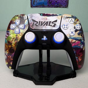 May include: A black wireless video game controller featuring a custom Marvel Rivals graphic skin on the front faceplate. The controller includes white analog sticks with blue LED lighting, a central PlayStation logo button, and standard directional and action buttons.