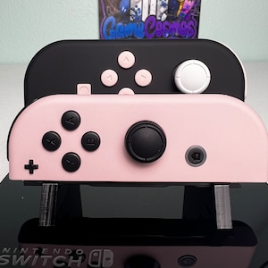 Może przedstawiać: Sterownik Nintendo Switch Joy-Con w kolorze różowym i czarnym z logo Nintendo Switch na dole. Sterownik ma czarną przednią płytę z różowymi przyciskami i białym joystickiem. Tylna część sterownika jest różowa.