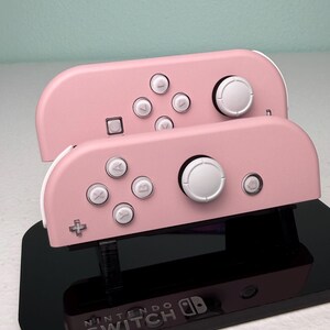 Może przedstawiać: Dwa r&oacute;żowe kontrolery Nintendo Switch Joy-Con z białymi przyciskami i czarną podstawką z logo Nintendo Switch.