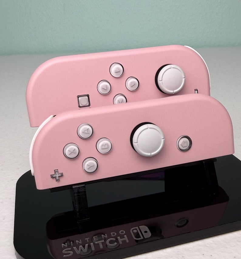 Może przedstawiać: Dwa r&oacute;żowe kontrolery Nintendo Switch Joy-Con z białymi przyciskami i czarną podstawką z logo Nintendo Switch.