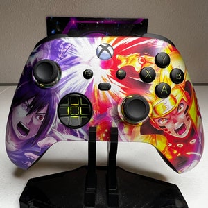 Könnte beinhalten: Ein weiß-schwarzer Xbox-Controller mit einem benutzerdefinierten Design, das die Charaktere Naruto und Sasuke aus der Anime-Serie zeigt. Der Controller hat einen schwarzen Ständer und gelbe Tasten.
