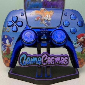 May include: A metallic blue video game controller featuring graphics of Sonic the Hedgehog, Tails, and Knuckles. The controller has blue buttons and joysticks, and it is displayed on a black stand with the text GameCosmos in purple and blue.