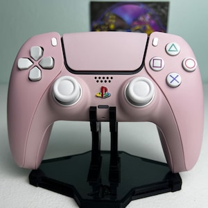 May include: A pink PlayStation 5 DualSense wireless controller with white buttons and a PlayStation logo on the front. The controller is on a black stand.