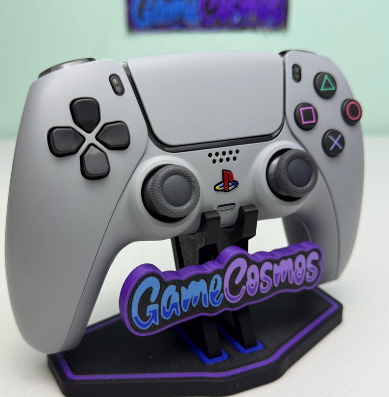 Puede incluir: Un mando gris de PlayStation 5 con botones y joysticks negros, apoyado en un soporte negro y morado. El soporte presenta el texto "Game Cosmos" en azul y morado. El logotipo de PlayStation es visible.