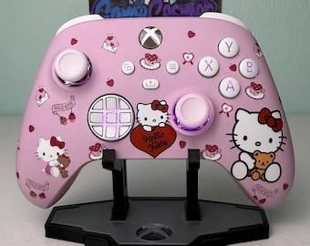 May include: A light pink wireless video game controller featuring Hello Kitty graphics, including a large Hello Kitty character holding a teddy bear, smaller icons of envelopes, cherries, and teacups, and the text Hello Kitty on a red heart.