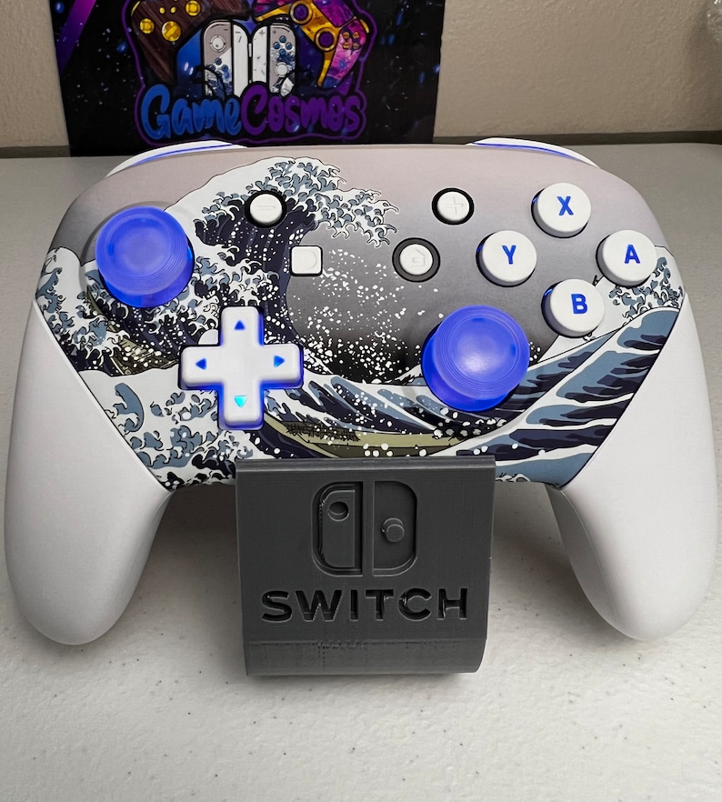 Puede incluir: Un mando Nintendo Switch blanco con un dise&ntilde;o personalizado que presenta una impresi&oacute;n de olas japonesas. El mando tiene botones azules y una empu&ntilde;adura gris. El mando est&aacute; conectado a un soporte gris con el logotipo de Nintendo Switch y la palabra "SWITCH" impresa en &eacute;l.